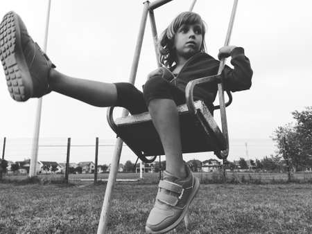 A 6-7 year old girl swinging on a swing against the background of a football field. Black and white monochrome image in retro or vintage style. Caucasian child.の写真素材