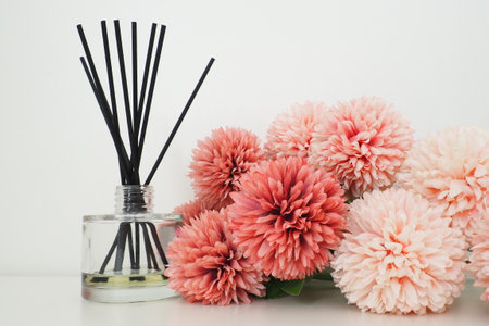 Composition with incense sticks, essential oil in a glass vase and pink terry artificial flowers in the interior of a white room. White background. Lots of flowers and scent sticksの写真素材