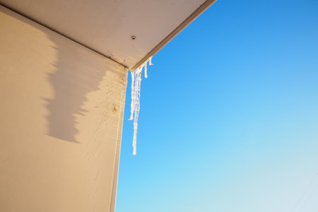 Long icicles dangle from the edge of a roof, capturing sunlight and glistening against a clear blue...