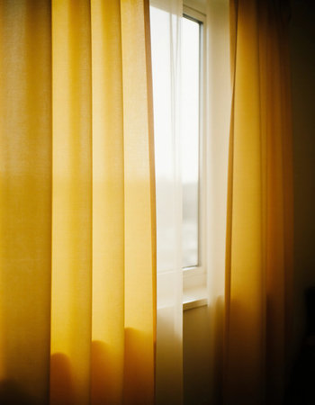 Bright yellow curtains drape elegantly by a window, softly filtering golden sunlight into the room in the morning and creating a cheerful, inviting atmosphere.の素材
