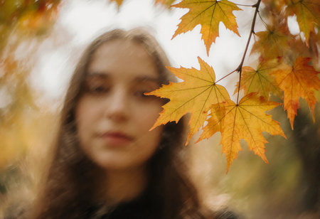A young woman stands in a serene setting, surrounded by bright autumn maple leaves. The scene is softly blurred, emphasizing the vibrant colors of fall foliage.の素材