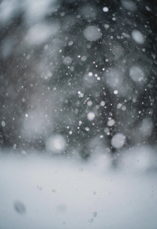 Snow covers the ground and falls from the sky in a winter setting. Tiny snowflakes float through the air while trees are partially visible in the background.の素材