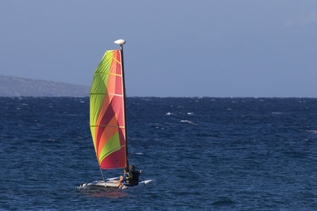 A couple sail on the deep blue Pacific on a catamaran with a brightly coloured sail.