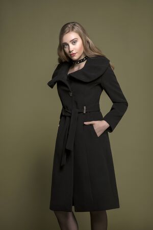 cute blonde model posing in trendy long black coat. olive green background.の写真素材