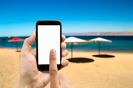 Hand holding smartphone with index finger touching on the beach