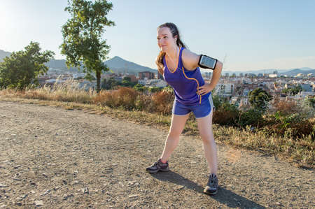 Fit runner woman taking a break while jogging. Young brunette female runner resting tired from her cardio workout relaxing while listening to music with a mp3 player and earphones.の写真素材