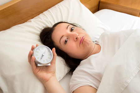 sad woman lying on the bed holding the clock and looking the camera