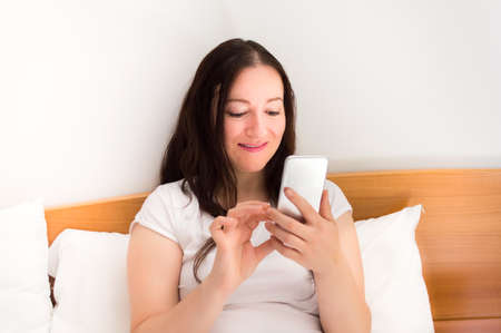 woman smiling reading a text message on the smartphone while lying in bedの写真素材