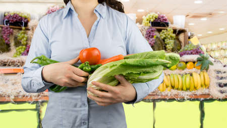 portrait of woman holding in her arms vegetables like lettuce tomatoes and peppers at the greengrocer on the backgroundの写真素材
