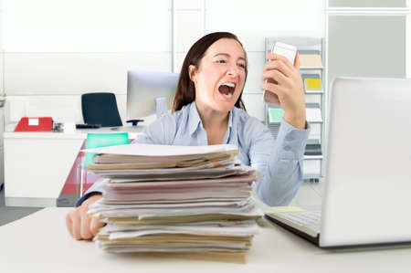 Angry female screaming into a cellphone sitting at a desk with a laptop and files of paperworkの写真素材