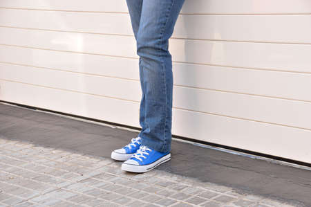 young woman with jeans and sneakers standing and leaning against a white metal door of a garageの写真素材
