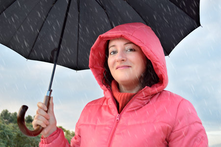 excited beautiful young woman wearing a pink anorak with hood holds a black umbrella in a fall rainy...