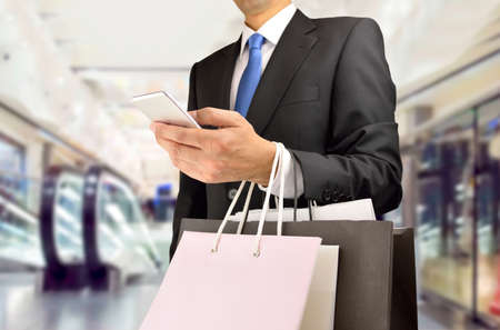 Cropped shot of shopper man holding the shopping bags an using the smart phone at the center mall. Concept of shopaholic man.の写真素材