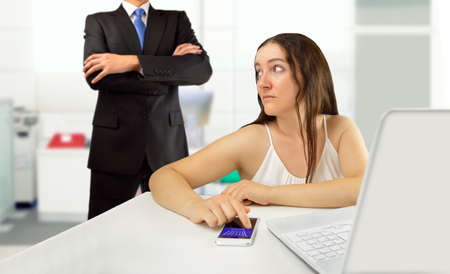 Lazy female employee playing games with her smartphone sitting in a desktop while her angry boss is watching at officeの写真素材