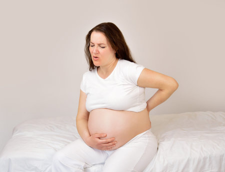 Pregnant woman suffering back ache sitting on a bed in the bedroom at homeの写真素材
