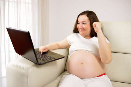 Excited pregnant woman buying on line and a laptop sitting on a couch in the living room in a house interiorの写真素材
