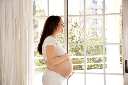 A beautiful pregnant woman holding her stomach affectionately thoughtfully looking out of a windowの写真素材