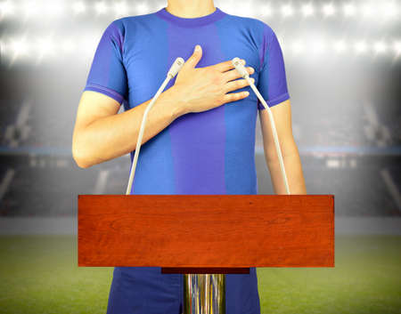 An empty podium and microphones with a young man and blue t-shirt with his hand on his heart and listening to his team's anthem at stadium background. Concept of love to a team or colors in sports.の写真素材