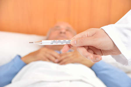 Hand of a doctor reading thermometer with a sick senior man lying in bed at hospitalの写真素材