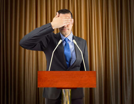 Portrait of an businessman covering his eyes with microphones over curtains in backgroundの写真素材