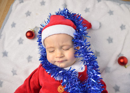 A cute newborn baby in white clothes sleeping in a basket on the floor against a twinkling Christmas...