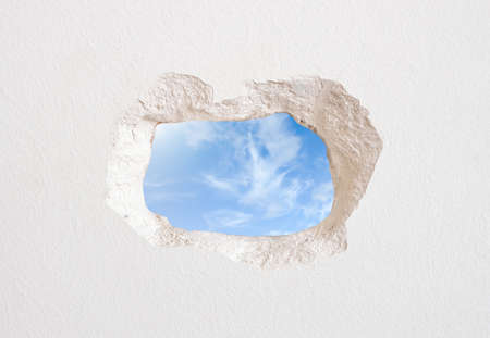 White wall with a hole that is observed the exterior of the building with clouds in the skyの写真素材