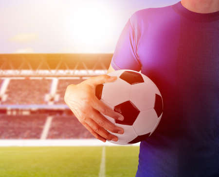 Cropped image of a young man holding a soccer ball with his hand wearing red tshirt and copy space at football stadium with a background sunsetの写真素材