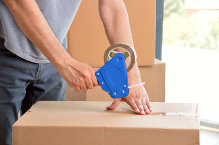 Shot of an unidentifiable young man closing a cardboard box with tape at homeの写真素材