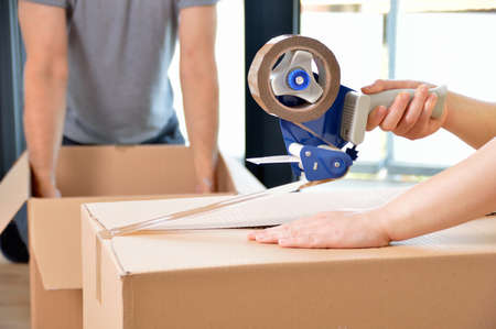 Close up of a couple moving home packing boxes and sealing with tape sitting on the floorの写真素材