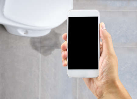 A hand holding a smartphone to call the professional to repair a water leak in the toiletの写真素材
