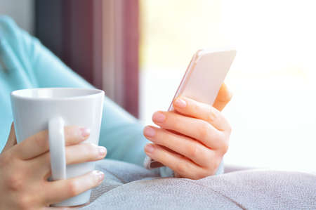 Woman hands holding a black mobile phone in a coffee break sitting in the living room with a window in the background and a warm lightの写真素材