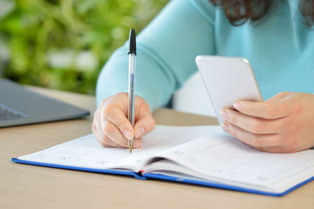 Close up of woman hand writing in agenda consulting a mobile phone on a desk at home or officeの写真素材