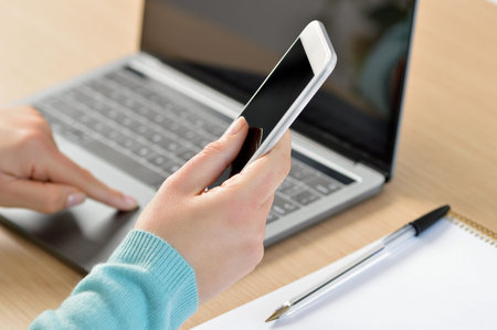 Close up of a woman hands using a smart phone and a laptop on a desktop at home or office