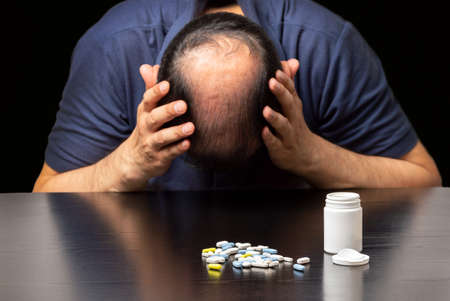 Close up of a depressed bald man next to a lot of pills on a table on a dark backgroundの写真素材