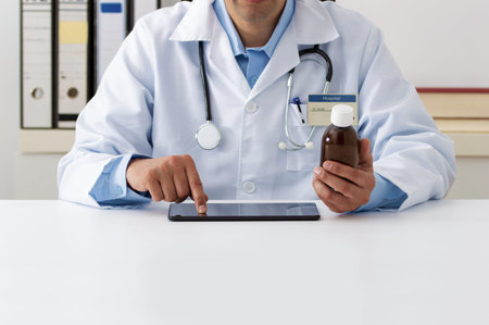 Photo of a young doctor consulting a tablet for a prescription in his office with a bottle of ...