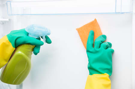 Close-up of woman's hand wearing yellow gloves cleaning open refrigerator with spray bottle and spongeの写真素材