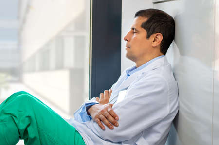 Cropped shot of an handsome young male doctor looking exhausted while leaning against a wall in her hospitalの写真素材