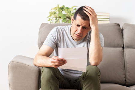 Furious man reading a letter sitting on a couch at homeの写真素材
