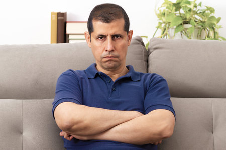 Front view portrait of an annoyed man looking at camera sitting on a couch in the living room at...