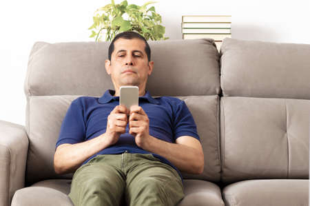 Bored man checking mobile phone sitting on a couch in the living room at homeの写真素材