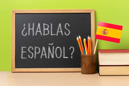 A chalkboard with the question hablas espanol? do you speak Spanish? written in Spanish, a pot with pencils and the flag of Spain, on a wooden desk with green backgroundの写真素材