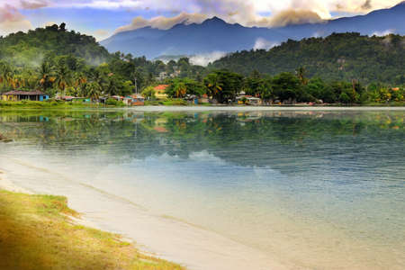 Beautiful landscape of small village at base of lush green mountains with dramatic sky aboe and reflective sea and sand belowの写真素材