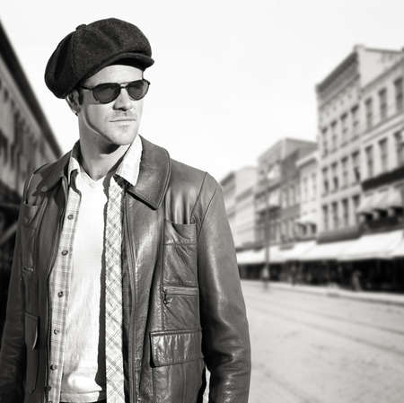 Sepia toned outdoor portrait of a cool young man in leather jacket, hat, and sunglasses against old town background with retro vintage feelingの写真素材