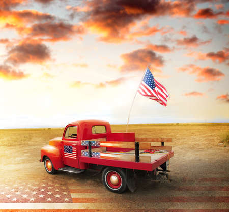 Red vintage pick up truck with American flag in wide open country side with dramatic sunset cloudscape and US flag on groundの写真素材