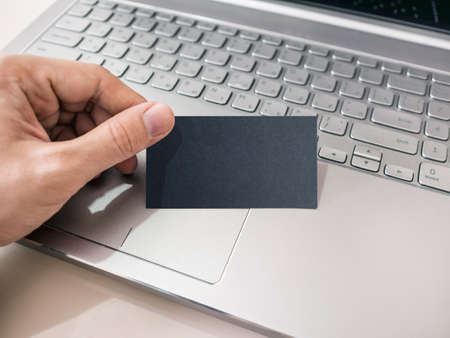 Man's hand holding blank Black Business Card. Modern Laptop on background, mockupの写真素材