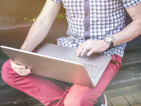 Young Man wearing in shirt sitting in city park and writing on his laptop, watch on wristの写真素材
