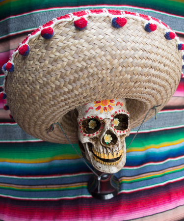 Decorated Dia De Los Muertos Skull wearing sombrero with colorful Mexican blanket in the background.