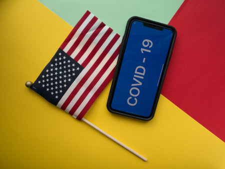 USA, March 16, 2020. USA flag and smartphone with the inscription COVID - 19 on a colored...