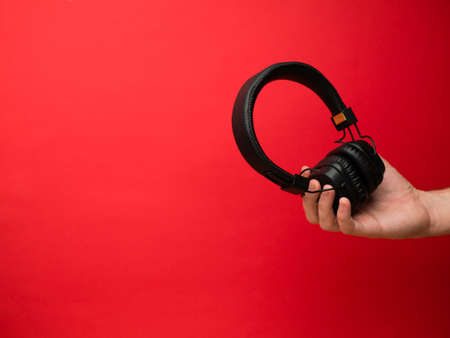 A young woman holds a pair of black wireless headphones in her hand. Hand of a girl with headphones on a red background. Musical concept.の写真素材