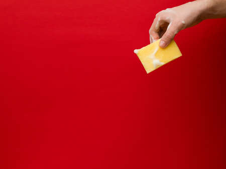 Male hand holding sponge with fingers. Washcloth covered with foam and bubbles. Copy space. Supplies for household. Domestic equipment for dishwashing concept. Isolated on red.の写真素材